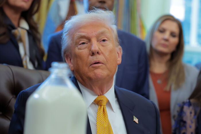 Donald Trump seated at a table with a large bottle of whole milk, surrounded by people in a formal setting.