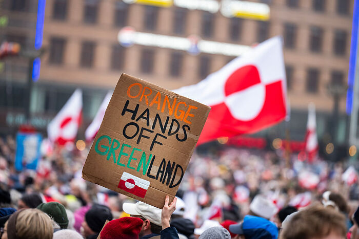 Protesters holding a cardboard sign in a crowd with Greenland flags representing a viral Greenland Reddit post message.