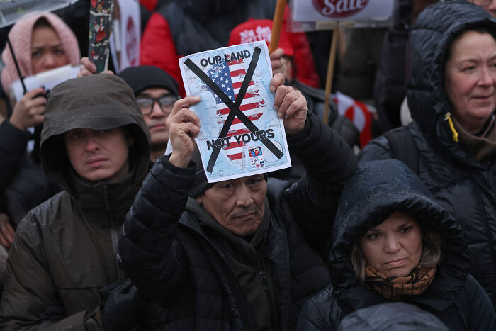 Crowd holding a crossed-out American flag sign protesting, illustrating the viral Greenland Reddit post telling Americans to stop apologizing.