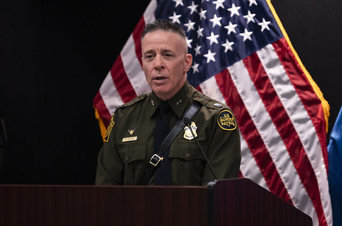 Border Patrol Chief Greg Bovino in uniform speaking at a podium with an American flag backdrop.