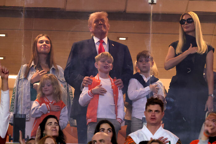 Donald Trump and family standing with hands over hearts during national anthem as crowd reacts with boos at sports event.