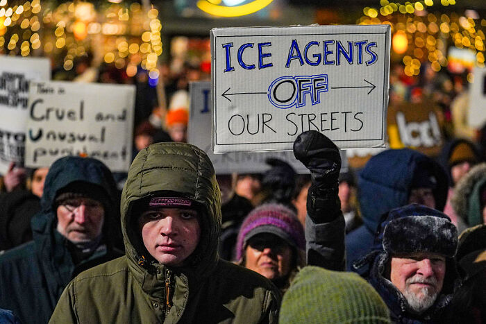 Protesters holding signs against ICE agents during a nighttime rally, reflecting backlash on ICE deployment plans.