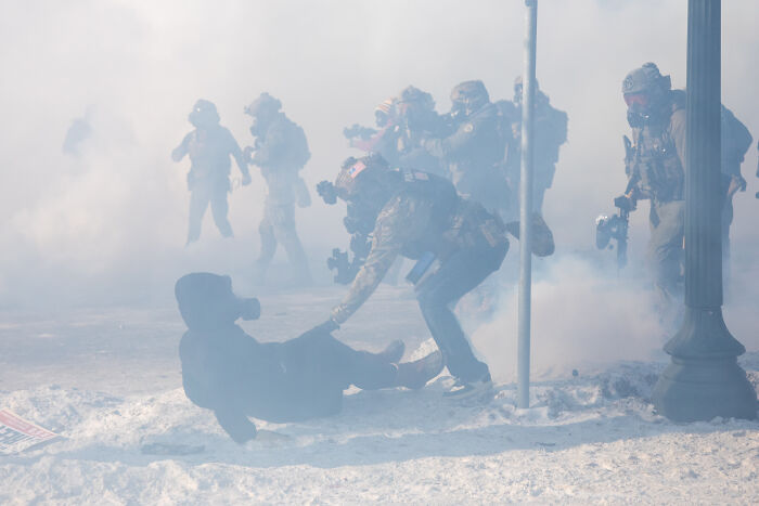 Police in tactical gear amid heavy smoke helping a person on the ground during a protest in winter.