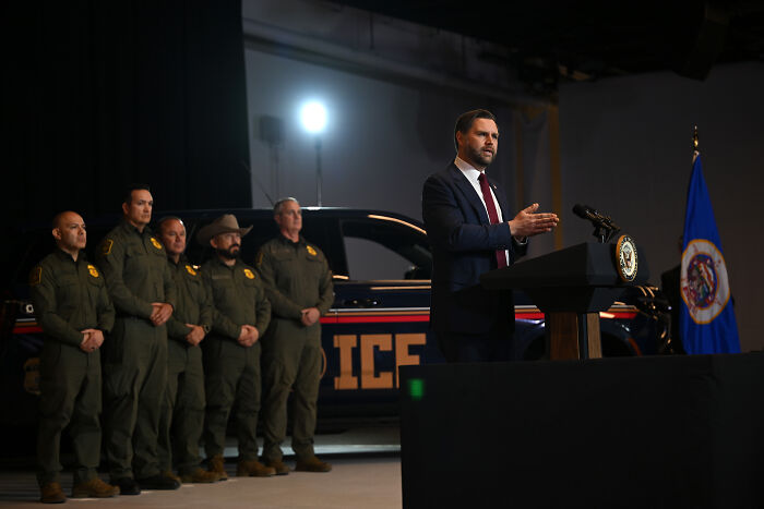 JD Vance speaking at a podium with law enforcement officers standing near an ICE vehicle during a public event.