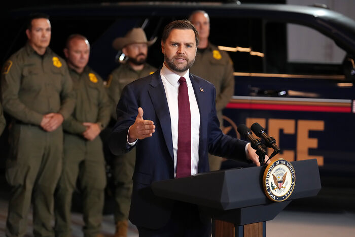JD Vance speaking at podium with law enforcement officers standing behind during a controversial hearing event.