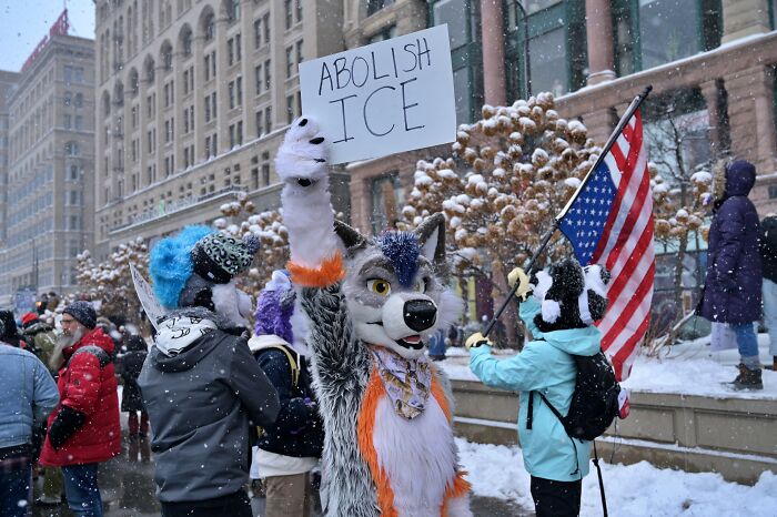 Protesters including a person in a wolf costume hold signs and an American flag voicing outrage at ICE in snowy urban setting.