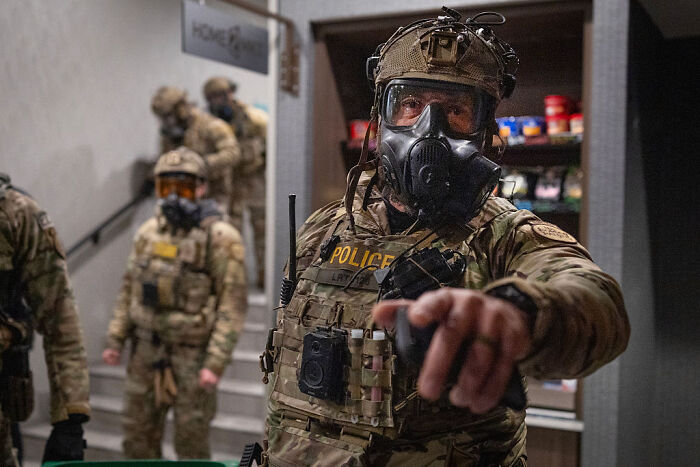 Police officer in tactical gear and gas mask pointing forward during an operation highlighting 2nd Amendment hypocrisy debate.