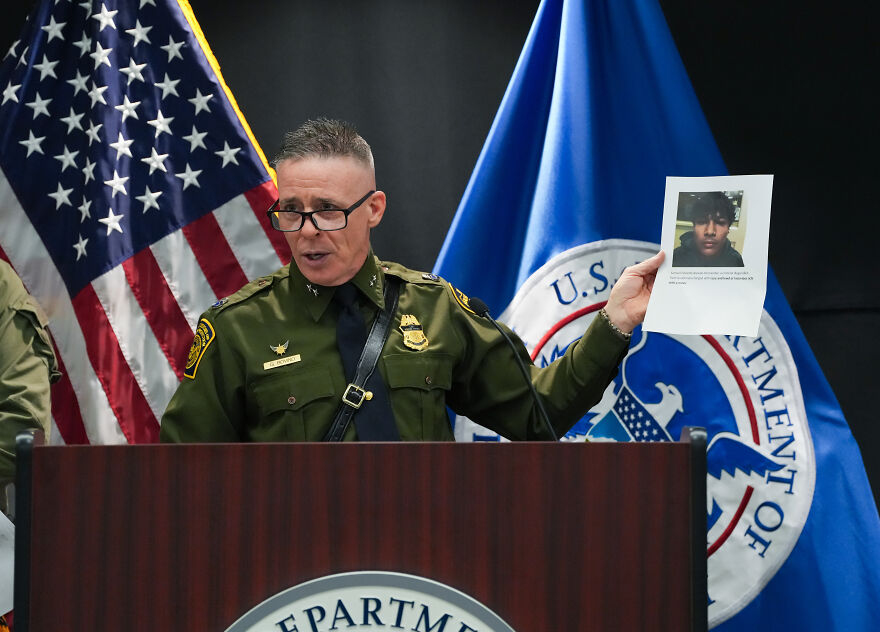 Border patrol agent speaking at a press conference with U.S. Department of Homeland Security and American flags behind.