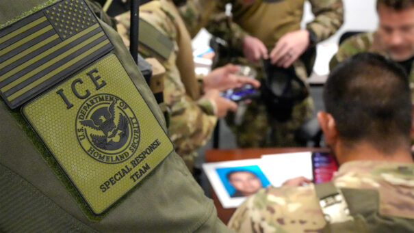 ICE agent patch on uniform with Homeland Security Special Response Team, officers gathered in background during briefing session