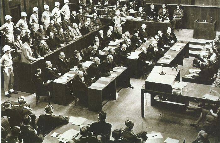 Courtroom scene with many people including guards and officials, illustrating a vital debate on an anti-ICE protest sign.
