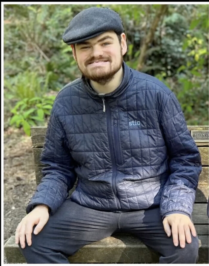 Young man sitting on a bench outdoors, wearing a quilted jacket and a cap, related to artist mom family tragedy news.