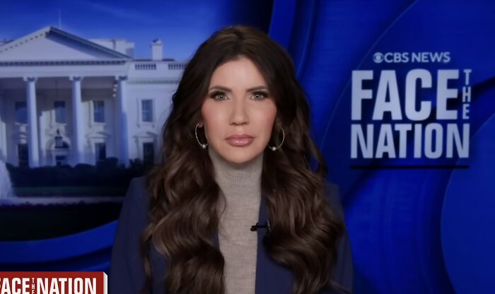 Kristi Noem on CBS News Face the Nation, wearing hoop earrings and a blazer with the White House backdrop behind her.