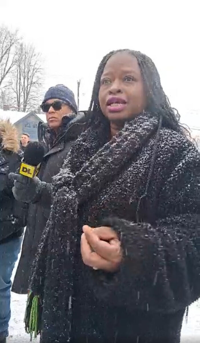 Woman speaking to media outside Minnesota church during protest on ICE pastor, snow falling on coats and scarves.
