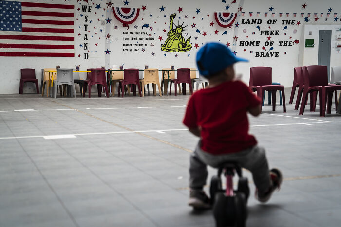 Child wearing a blue cap riding a tricycle inside a Texas ICE facility with patriotic decorations on the walls.