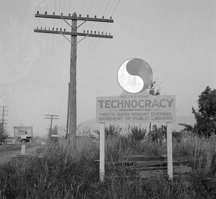 Black and white photo of a technocracy sign by a roadside with power lines and grassy fields in the background.