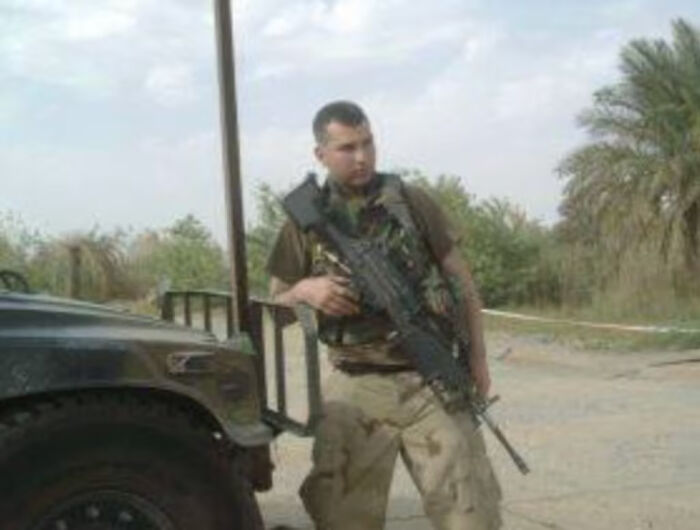 ICE agent in tactical gear holding a rifle, standing outdoors near a military vehicle in a desert-like area.