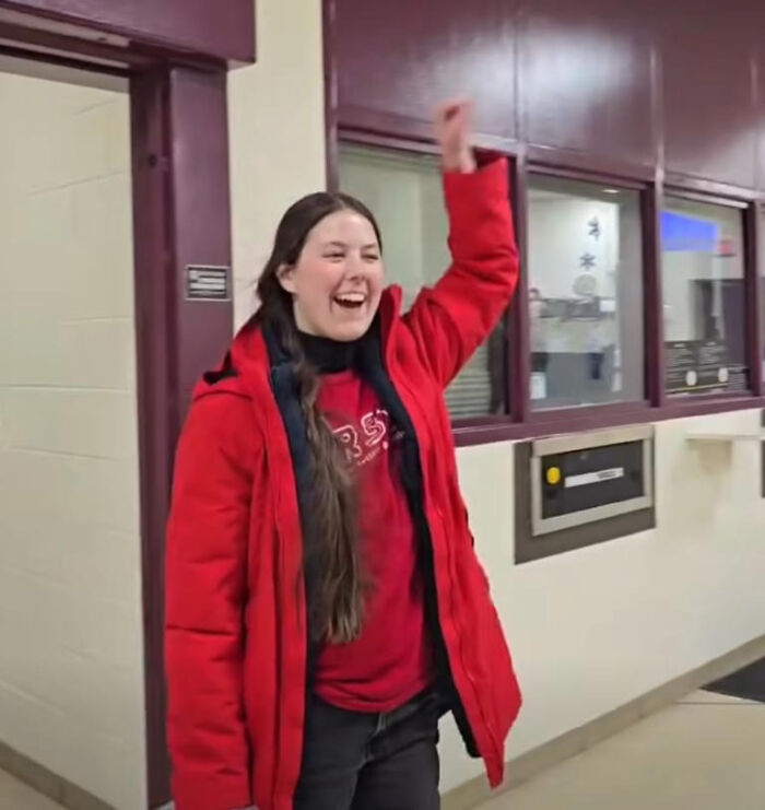 Preschool teacher in red coat smiling and raising an arm in a school or institutional hallway.