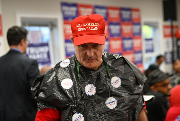 Man wearing Make America Great Again hat and multiple Trump buttons, looking sad and confused at event.