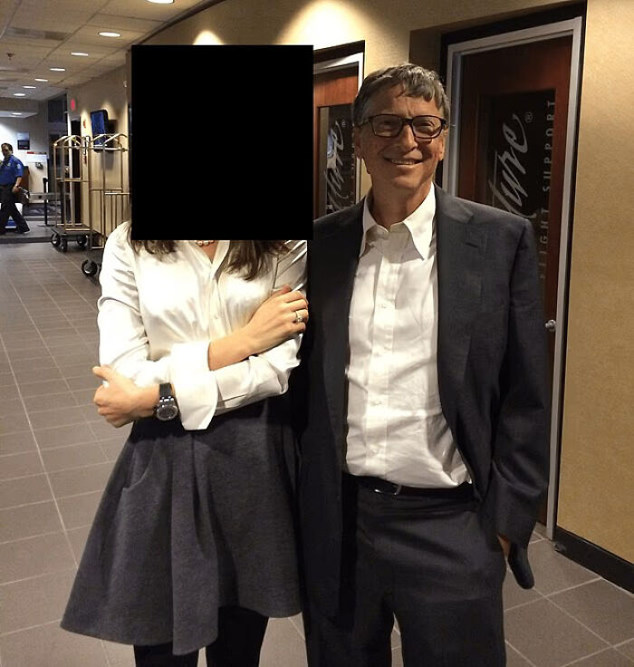 Bill Gates standing indoors in a suit smiling next to a woman with face obscured in a hallway setting.