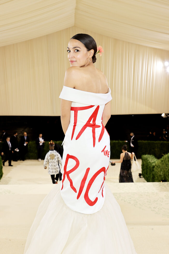 AOC posing in a white gown with red lettering at an event, signaling possible White House run in 2028.