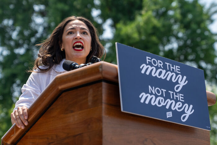AOC speaking passionately at a podium outdoors with a sign reading for the many not the money.