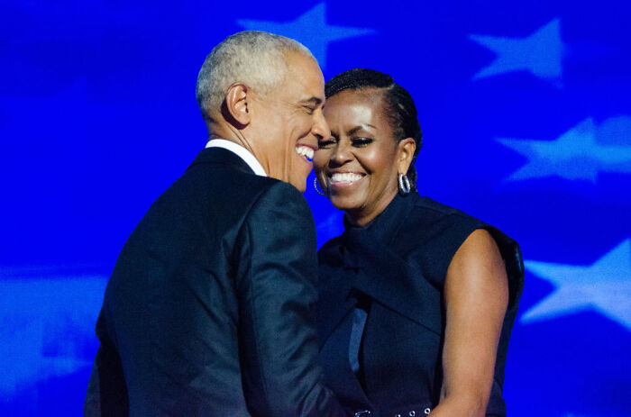 Barack and Michelle Obama sharing a joyful moment on stage with blue star-patterned background.
