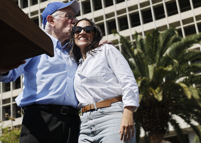 AOC smiling in sunglasses and white shirt with man in blue shirt and hat, signaling a White House run in 2028.