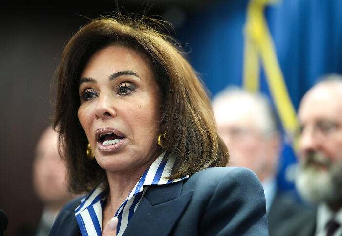 Woman speaking at a press conference with blurred individuals and flags in the background discussing grand jury blocking DOJ charges.