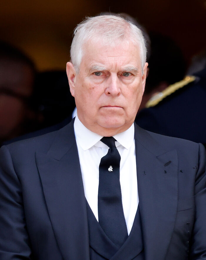 Elderly man in a formal black suit and tie with a serious expression amid reputation and scandal concerns related to King Charles.