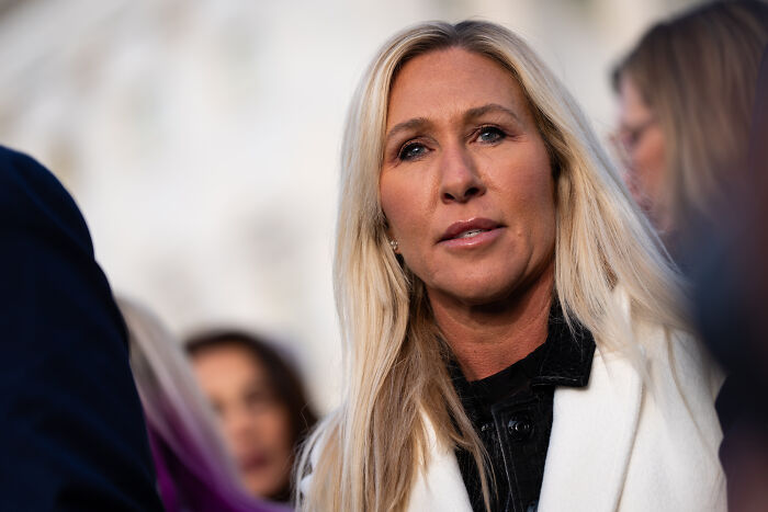 Marjorie Taylor Greene speaking to media outdoors, criticizing weak Republicans and labeling them cowards.