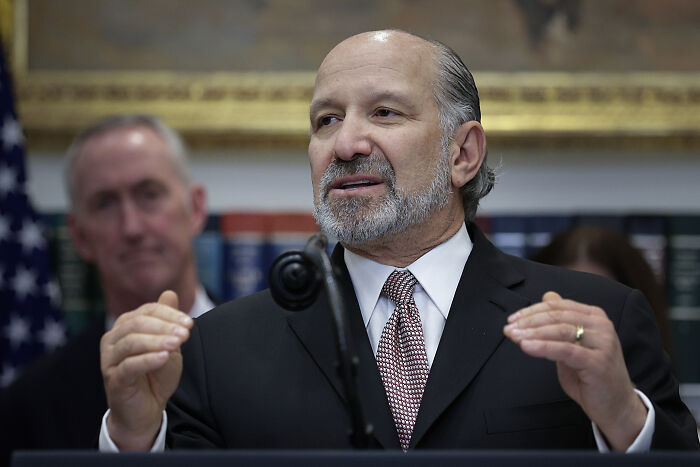 Man in suit speaking at podium during press event, with focus on Trump dodging Epstein survivor questions and reporter interaction.