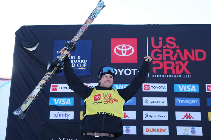 U.S. Olympic athlete celebrating at U.S. Grand Prix ski event with raised arms and skis, showcasing mixed emotions.
