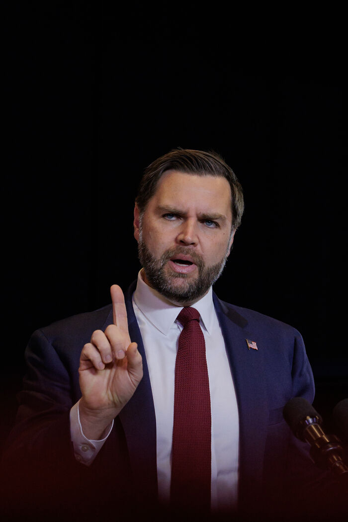 JD Vance speaking seriously at a podium, wearing a suit and red tie, making a gesture with his index finger raised.