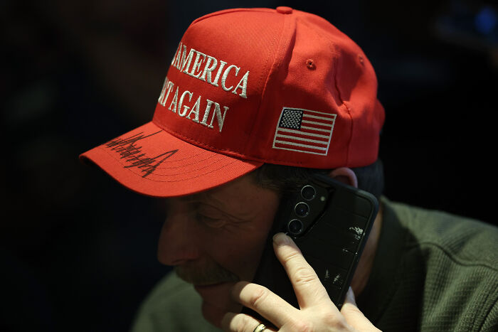 Man wearing red MAGA hat with American flag patch speaking on phone in low light, illustrating MAGA ideologies on dates.
