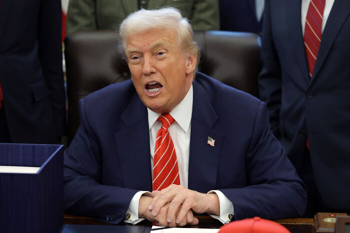 Man in suit and red striped tie speaking emphatically while seated at a desk, with others standing behind him.