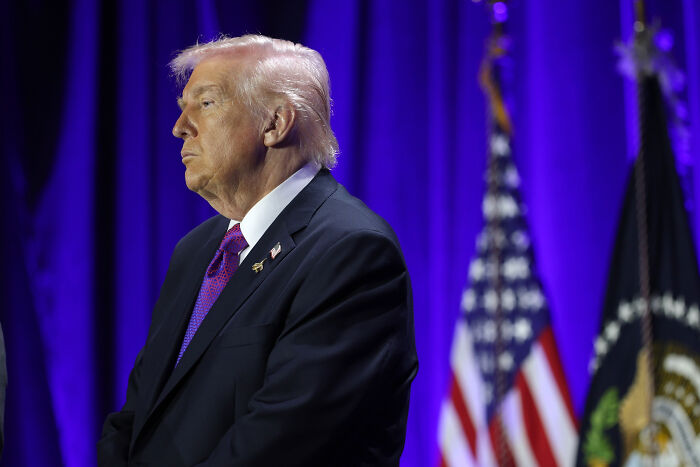 Donald Trump at National Prayer Breakfast, standing in front of American flags with a serious expression.