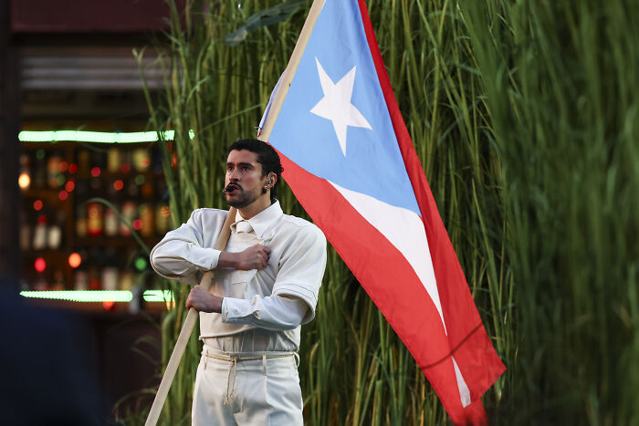 Man in vintage outfit holding Puerto Rican flag outdoors with tall grass in the background during an event.
