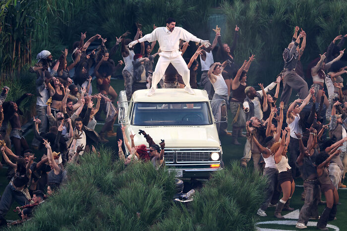 Man in white outfit dancing on van roof surrounded by crowd, highlighting Super Bowl party and Bad Bunny music scene.