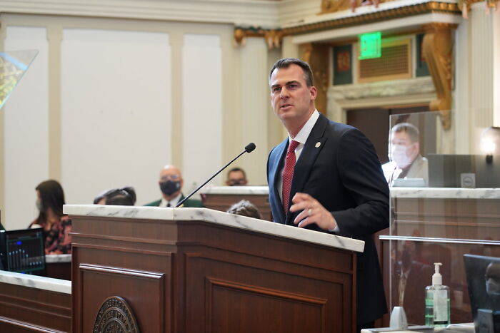 Man in a suit speaking at a podium in a government chamber during a dispute involving a RINO governor.