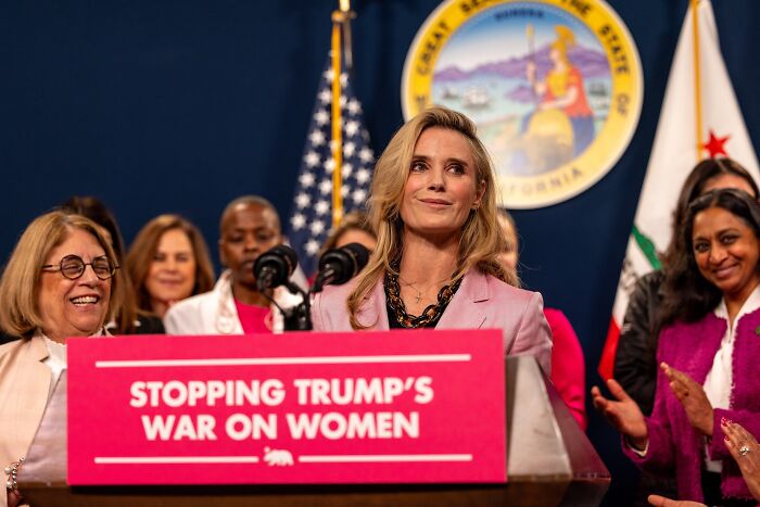 Jennifer Siebel Newsom speaking at a podium with a sign about stopping the war on women, addressing reporters.