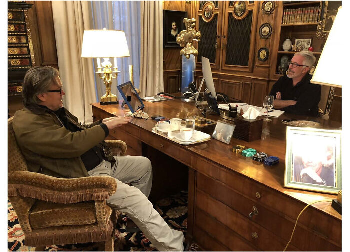 Two men in a wood-paneled room having a discussion at a large desk with lamps, referencing MAGA influencers and Epstein ties.