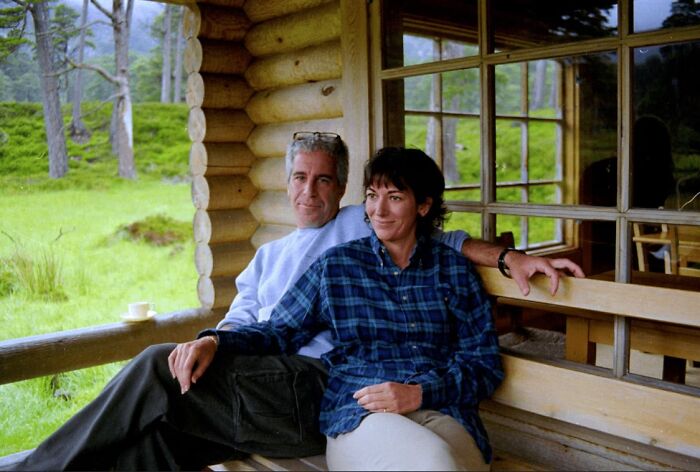 Couple sitting on a wooden porch bench outside a cabin in a green, forested area on a cloudy day.