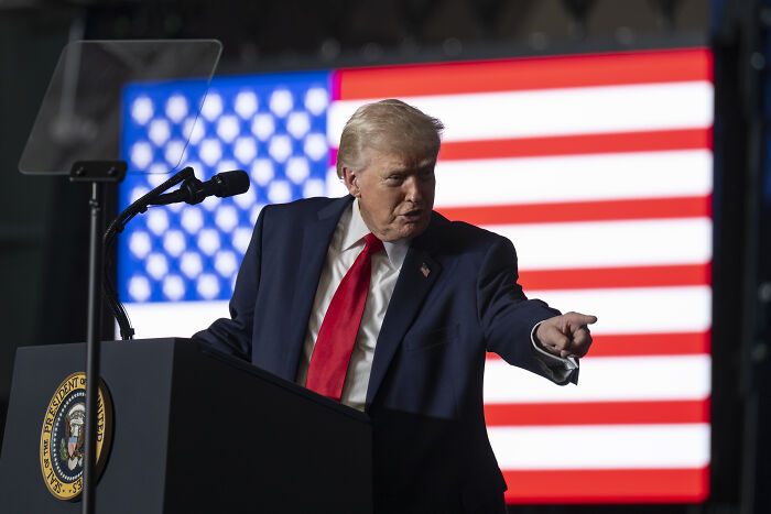 Donald Trump at a podium pointing, with an American flag backdrop, amid dispute involving RINO governor and Republican dinner.