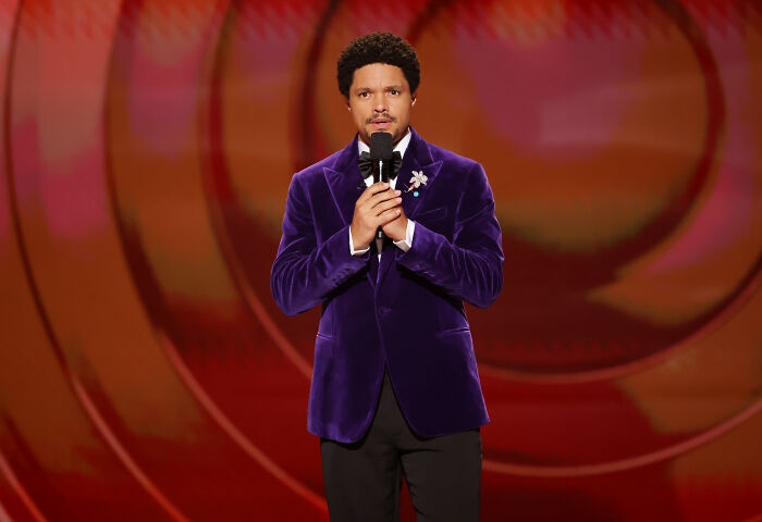 Trevor Noah in a purple velvet jacket holding a microphone, performing a comedy routine on stage.
