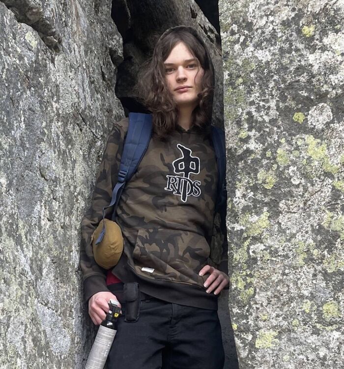 Young person in camo hoodie and backpack holding an object, framed by large rugged rocks, hinting at disturbing Canada school tragedy.