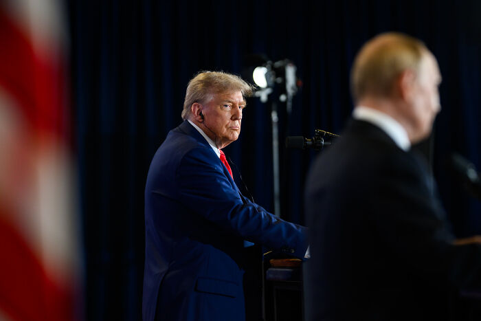 Former President Trump at a podium, listening during a political event, with a blurred figure in the foreground.
