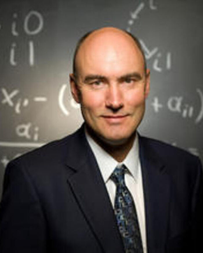 Harvard professor wearing a suit and tie standing in front of a chalkboard with mathematical equations in a classroom setting