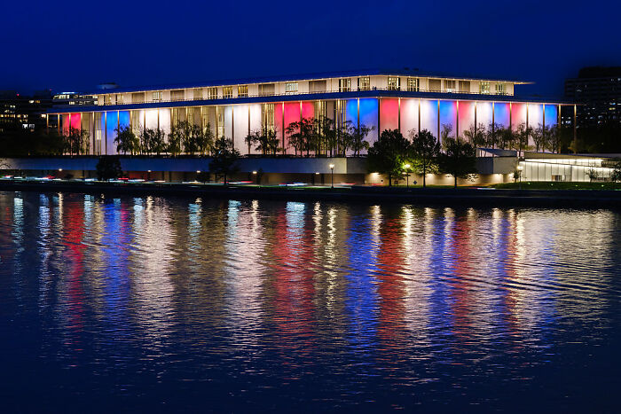 The Kennedy Center illuminated at night with red, white, and blue lights reflecting on the water nearby.