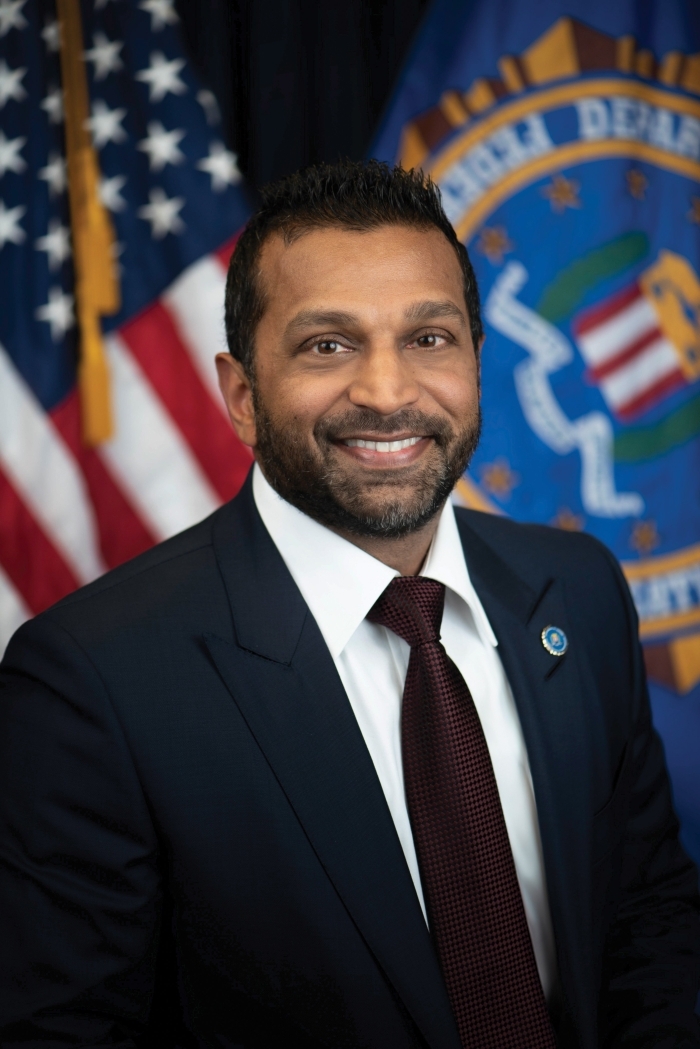 Patel, a Trump official, smiling in a dark suit and burgundy tie, with an American flag and seal in the background.