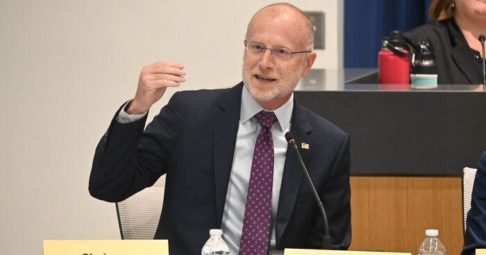 A man in a suit and tie, wearing glasses, speaking at a table with a microphone. He discusses Jimmy Kimmel.